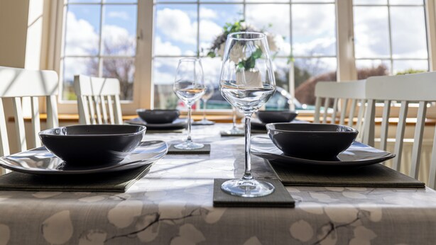 Une table à manger dressée avec des bols noirs, des verres à vin et un vase rempli de fleurs.