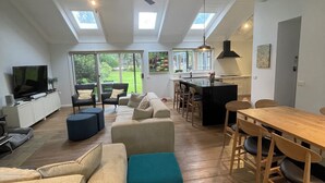 Un vaste espace de vie comprenant un canapé, un pouf, une table à manger et des chaises. On y trouve également un téléviseur, un ventilateur de plafond et des fenêtres de toit.