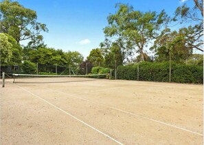 Ein Tennisplatz im Freien mit Netz, umgeben von Bäumen und einer Hecke.