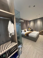Quarto de hotel moderno com guarda-roupa de madeira, cama com cabeceira, mesa de cabeceira com abajur e uma janela grande com cortinas.