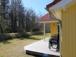 Eine Holzterrasse mit Stühlen, einem überdachten Bereich und einem gelben Gebäude mit rotem Dach.
