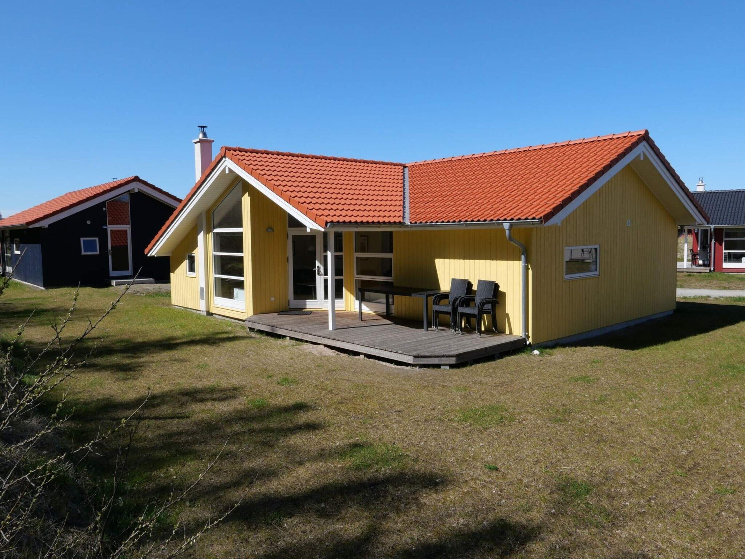 A yellow building with a red roof, a covered patio with chairs, and a grassy yard.