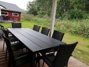 Eine Holzterrasse mit Tisch und Stühlen, umgeben von Grünpflanzen und einem Haus im Hintergrund.