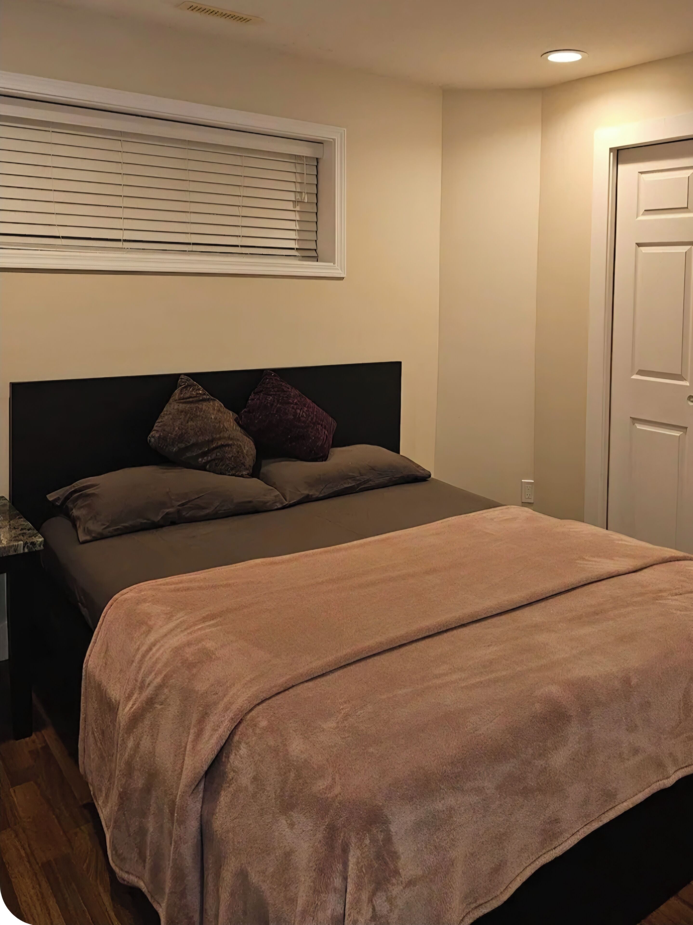 Une chambre à coucher avec un lit, une fenêtre avec des stores, une porte et un luminaire au plafond.