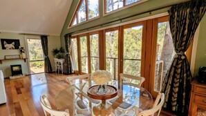 A dining area with a round glass table, white chairs, and a view of trees through large windows. - Bickley Tree Stay (Bickley)