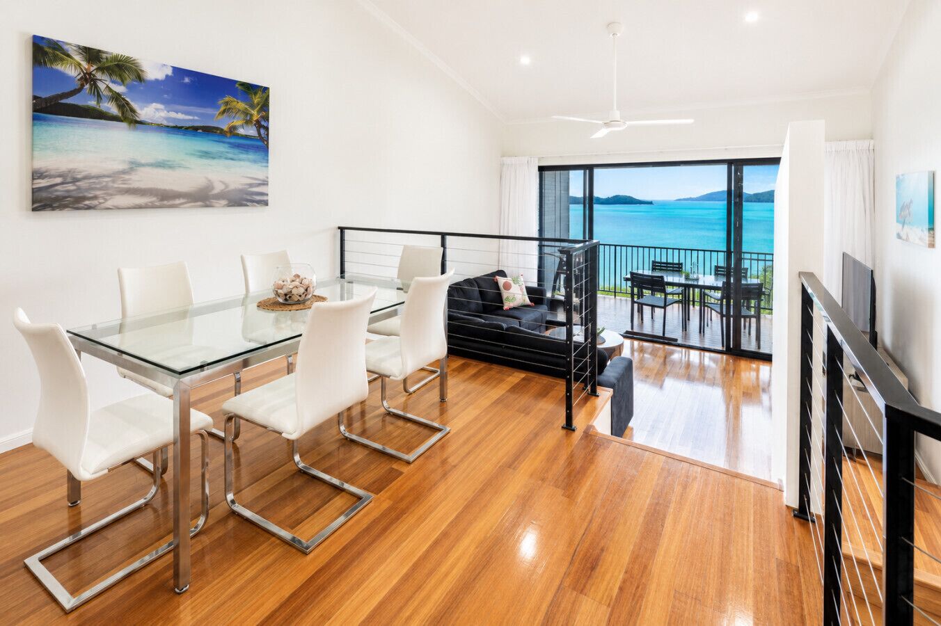 Een moderne woonkamer met een glazen eettafel, witte stoelen, een zwarte bank en een balkon met uitzicht op zee.