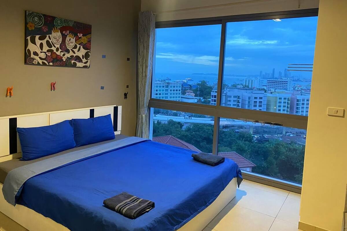 A bedroom with a large bed, a city view, and a painting on the wall.