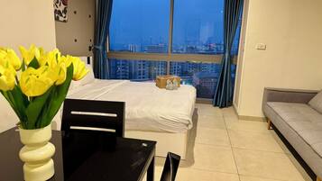 A modern hotel room with a large window, a bed, a dining table, and a sofa.