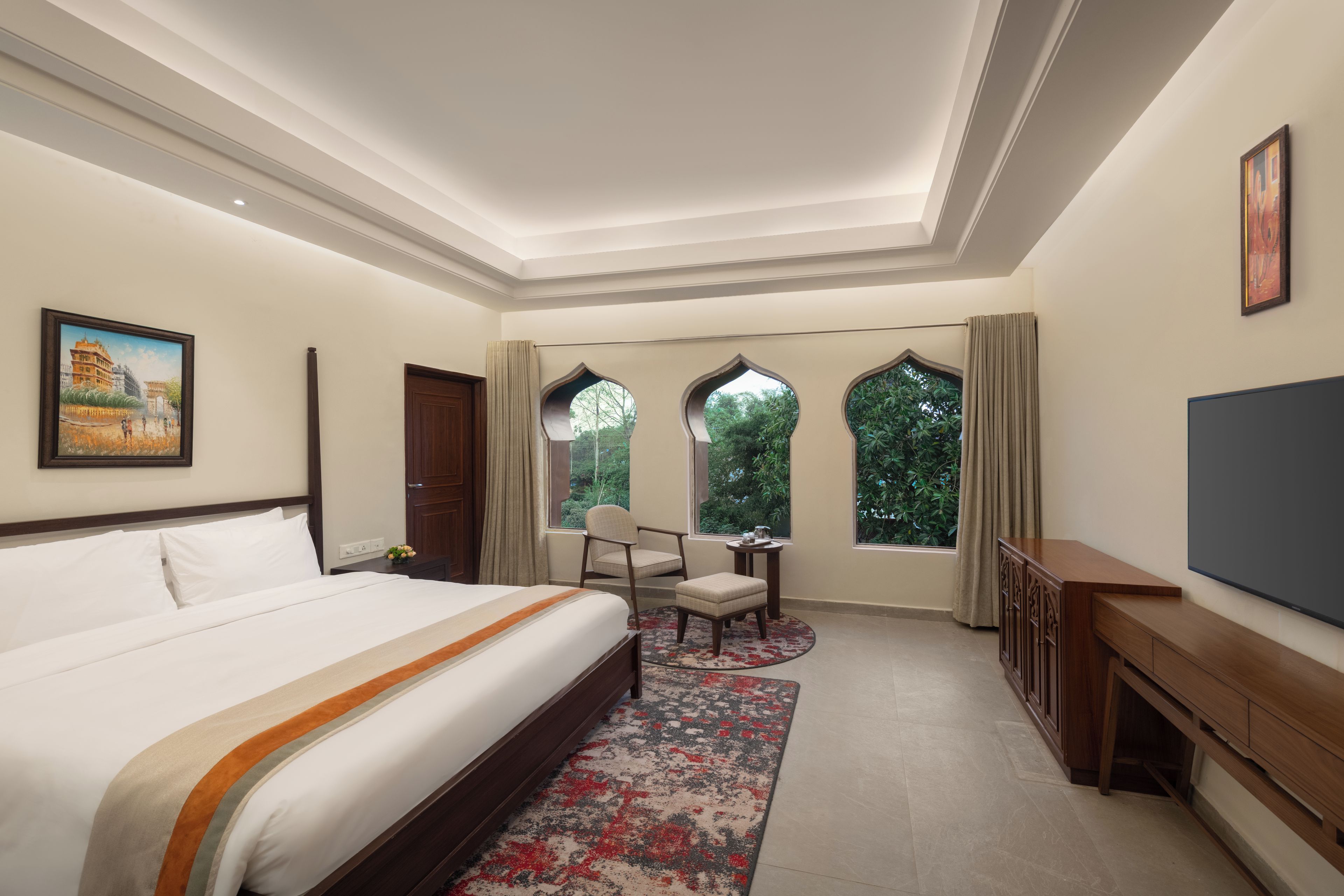 A hotel room with a large bed, a desk, a chair, a TV, and a window with a view of greenery.