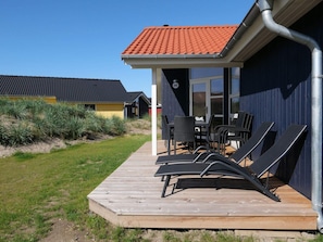 Eine Holzterrasse mit Liegestühlen, einem Tisch und einem überdachten Sitzbereich mit rotem Ziegeldach.