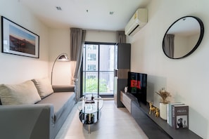 A modern living room with a sofa, a glass coffee table, a flat-screen TV, a mounted mirror, and a bookshelf with books and decorative items. - 5 Star Sukhumvit 1BR apt Pool, Gym, Sauna Thong lo (Krung Thep Maha Nakhon)