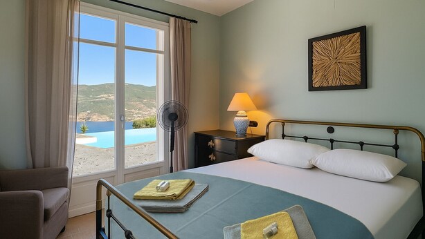 A bedroom with a bed, bedside table, lamp, and a view of a body of water through large windows. - Brand-New Villa 'Maison Magnolia', Infinity Pool & Sea View above Skopelos Town (Skopelos)