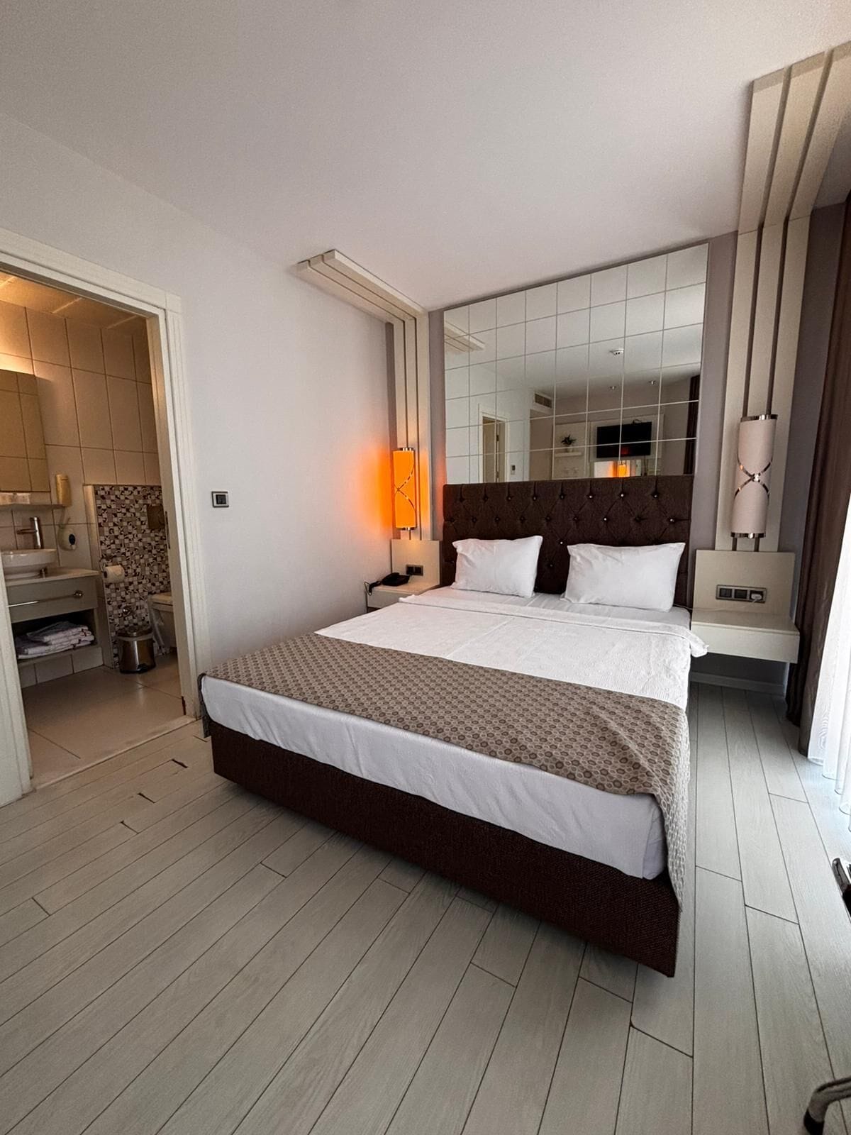Une chambre d’hôtel moderne avec un grand lit, une table de chevet, un miroir et une salle de bain visible à travers une porte ouverte.