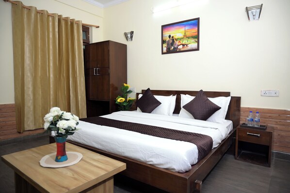 A hotel room with a bed, a nightstand, a wooden table with a vase of flowers, a wardrobe, and a painting on the wall.