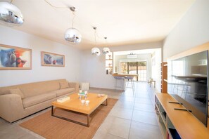 A modern living room with a sofa, coffee table, and TV. There are paintings on the wall and a dining area in the background. - Villa Marathe Lux | Akrotiri, Chania | Sea Views | Private Pool (Crete)