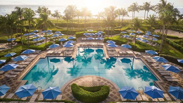 Ein Pool in einem Resort mit mehreren Liegestühlen und Sonnenschirmen, umgeben von Palmen und Blick auf den Ozean.