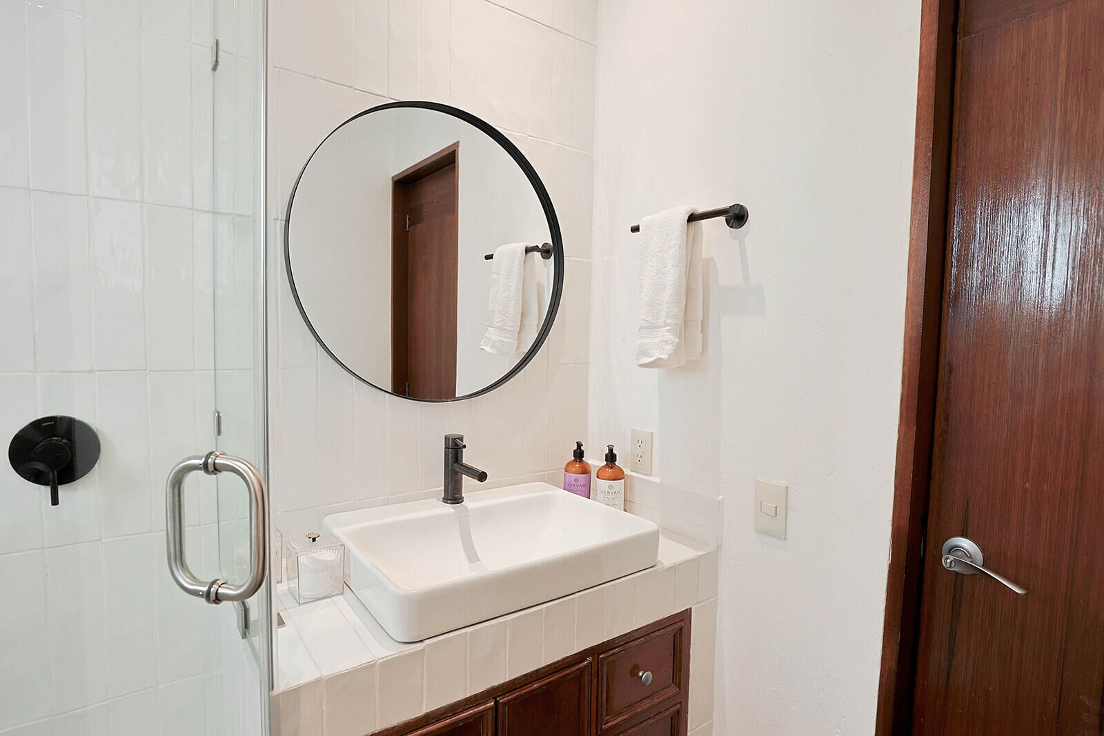 Un baño con un lavamanos blanco, un espejo redondo, un perchero para toallas y una puerta de madera.