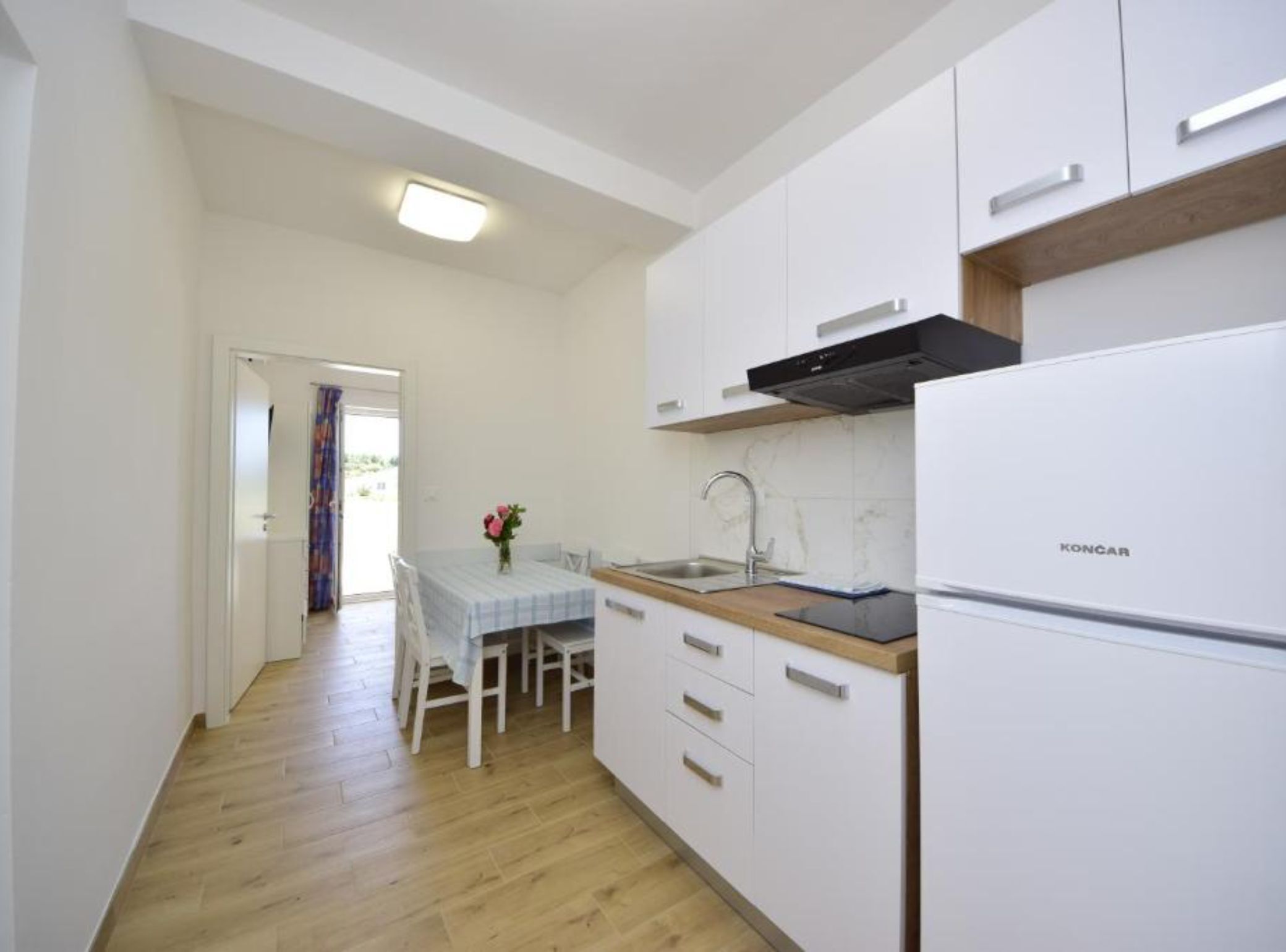 Una cucina compatta con mobili bianchi, un frigorifero Koncar, un piano di lavoro in legno e un tavolo con sedie.