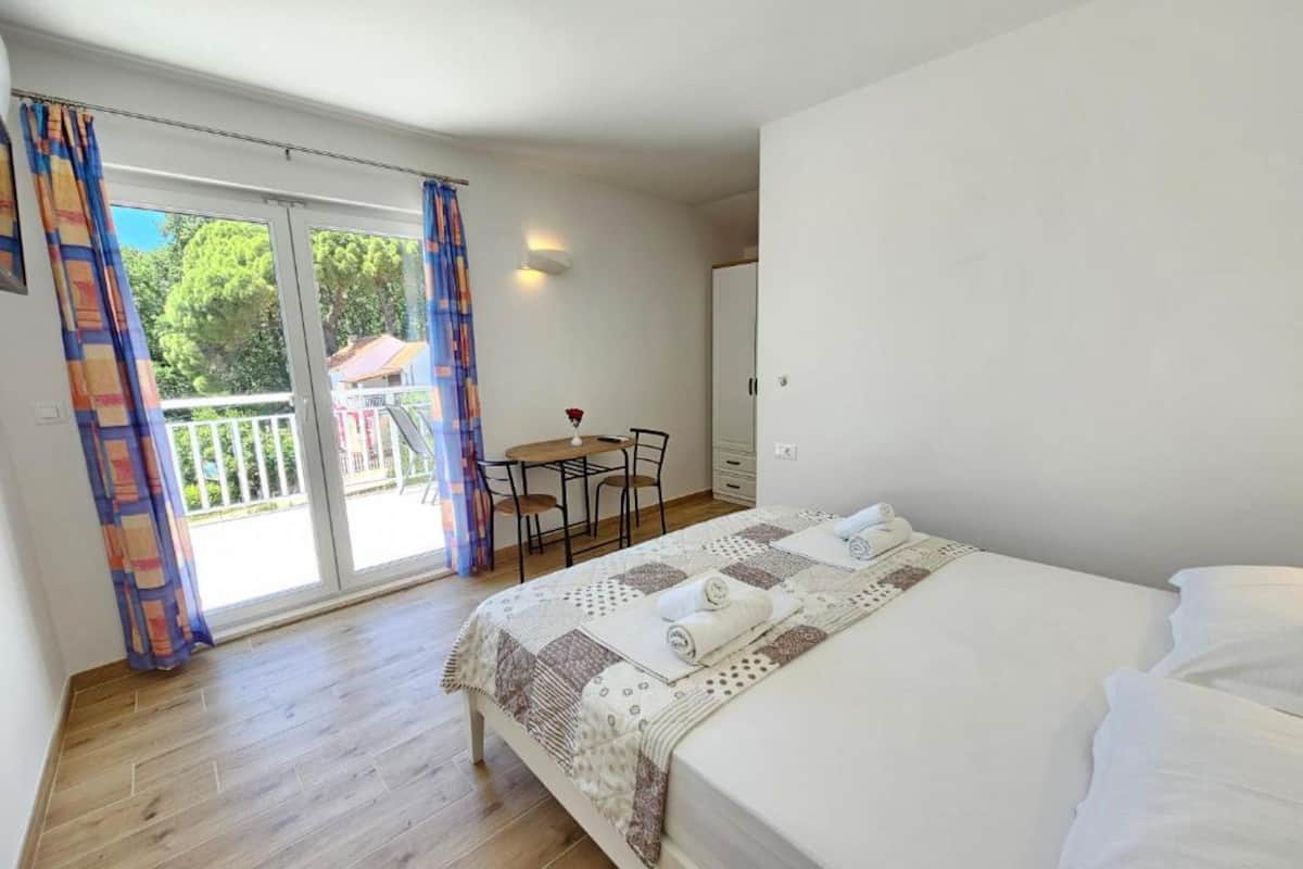 A bedroom with a bed, a small table, and a chair, a balcony with curtains, and a view of greenery outside.