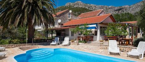 Un espace aménagé au bord de la piscine avec des chaises longues, un bâtiment doté d’un toit recouvert de tuiles rouges et un paysage montagneux en arrière-plan.