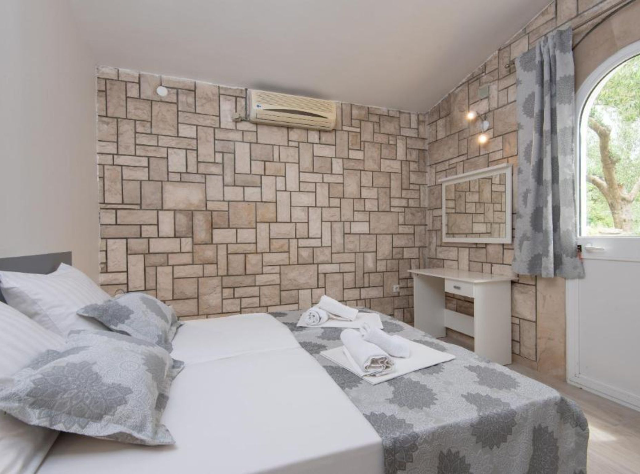 A bedroom with a brick-patterned wall, a bed with a patterned bedspread, a bedside table with a mirror, and a window with curtains.
