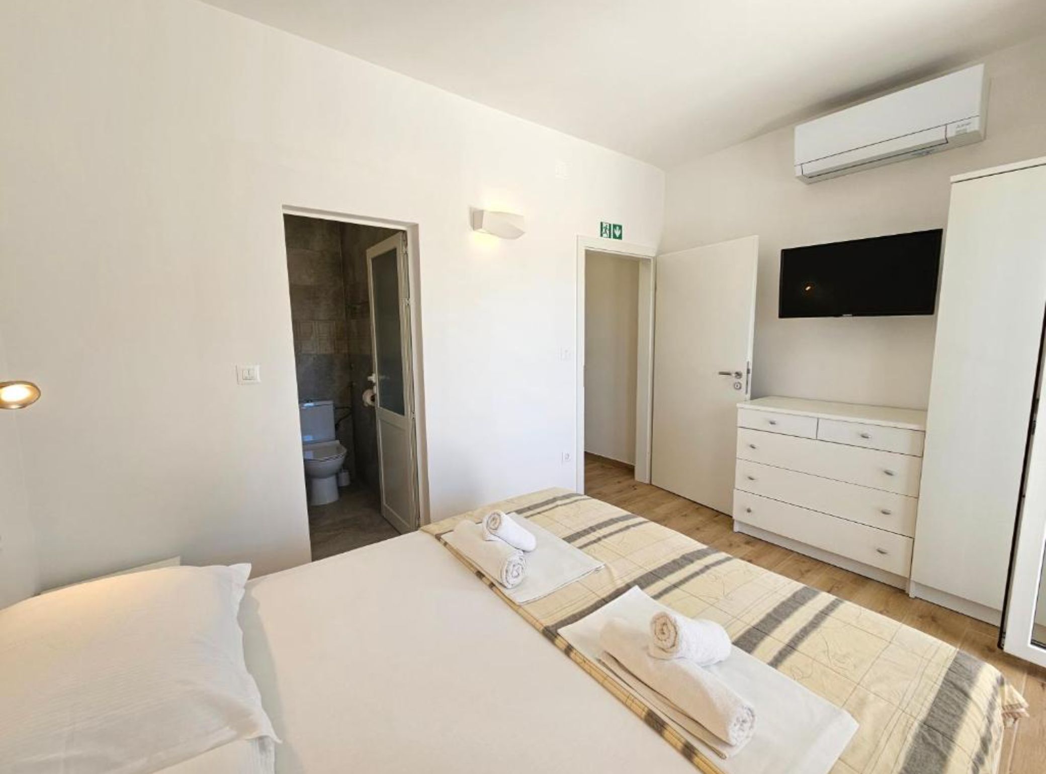 A hotel room with a bed, a bathroom, a wardrobe, a flat-screen TV, and an air conditioning unit.