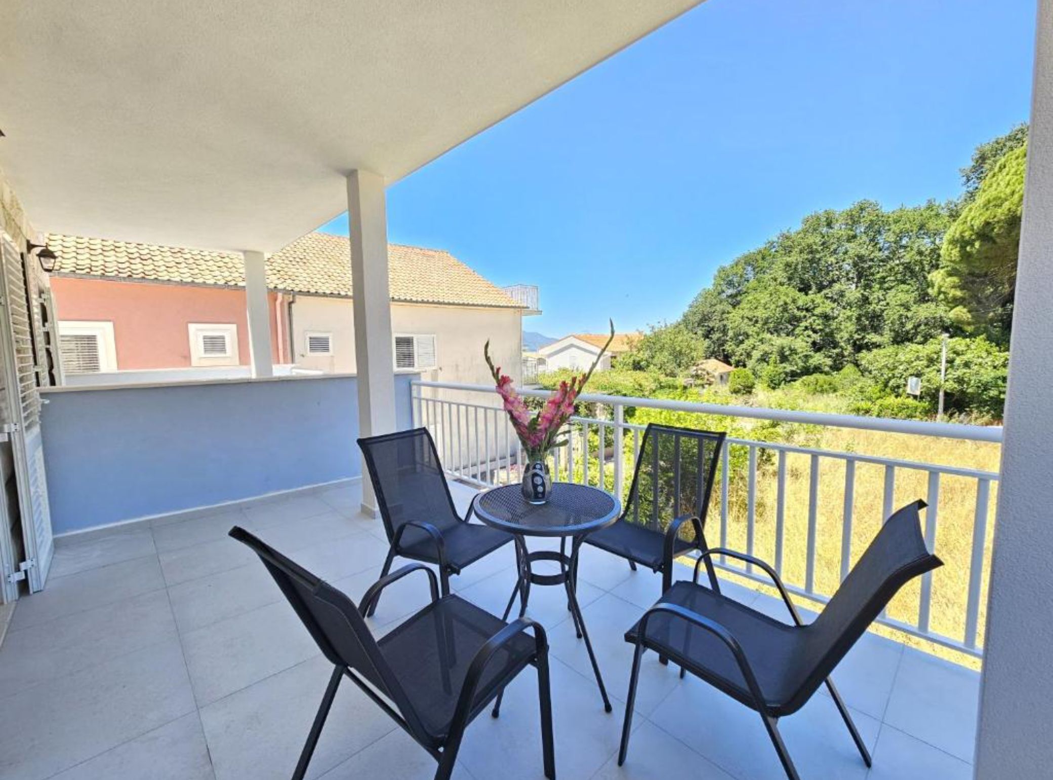 Un balcon avec une table et des chaises, un vase rempli de fleurs et une vue sur des immeubles et des arbres.