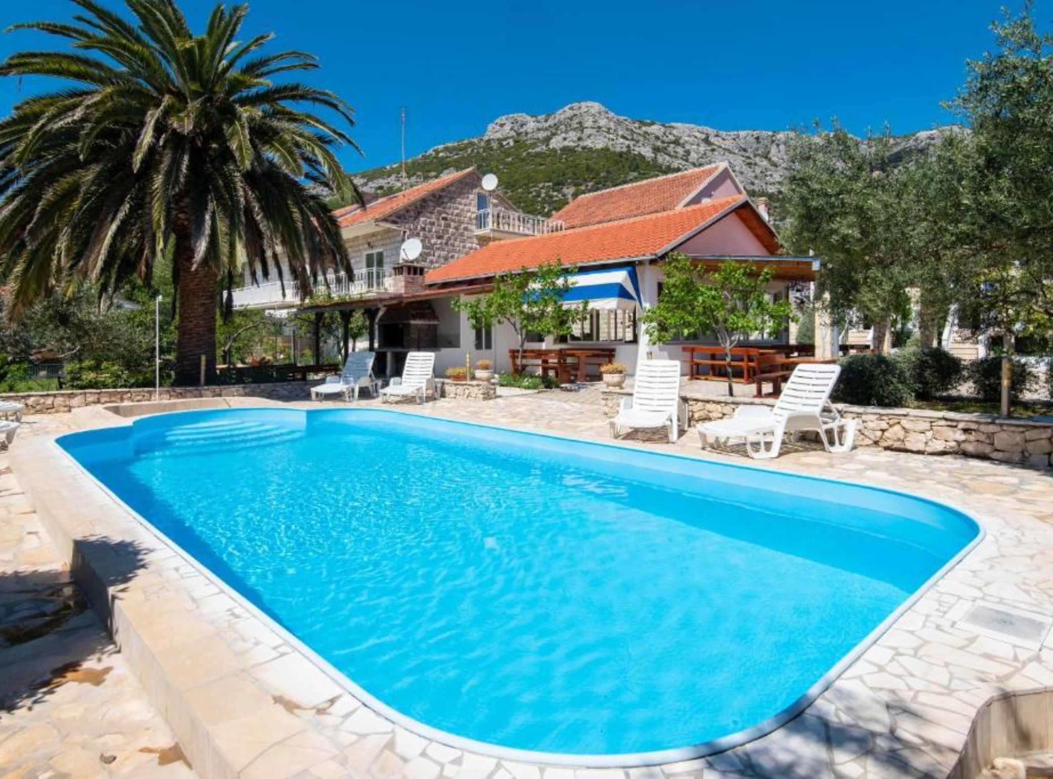 Una piscina con sillas de descanso, un edificio de piedra con techo rojo y una montaña al fondo.