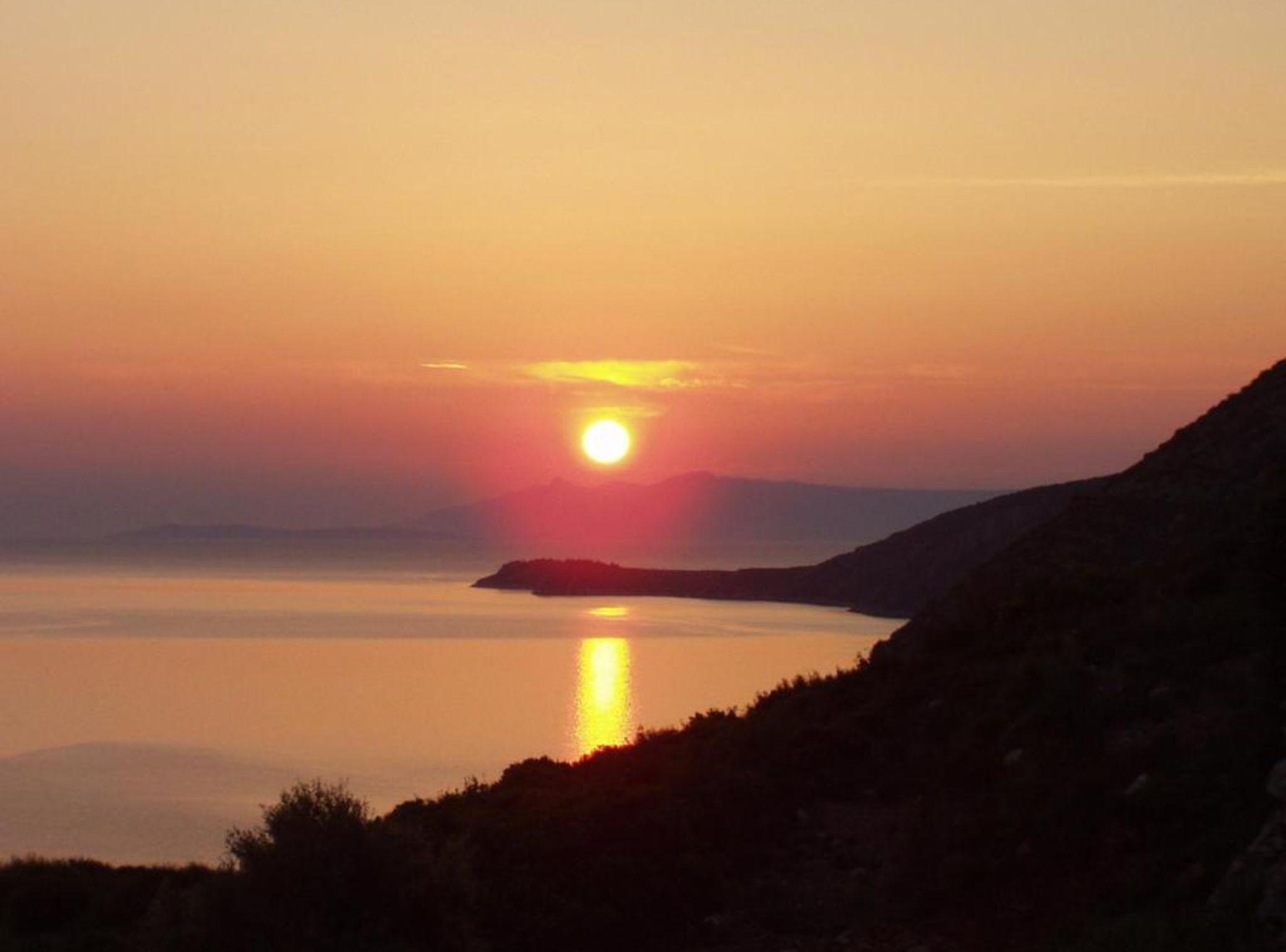 Un coucher de soleil sur une étendue d’eau calme, avec la silhouette d’une chaîne de montagnes à l’horizon.