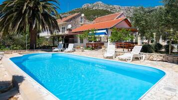 A swimming pool with lounge chairs, a stone building with a red roof, and a mountain in the background.