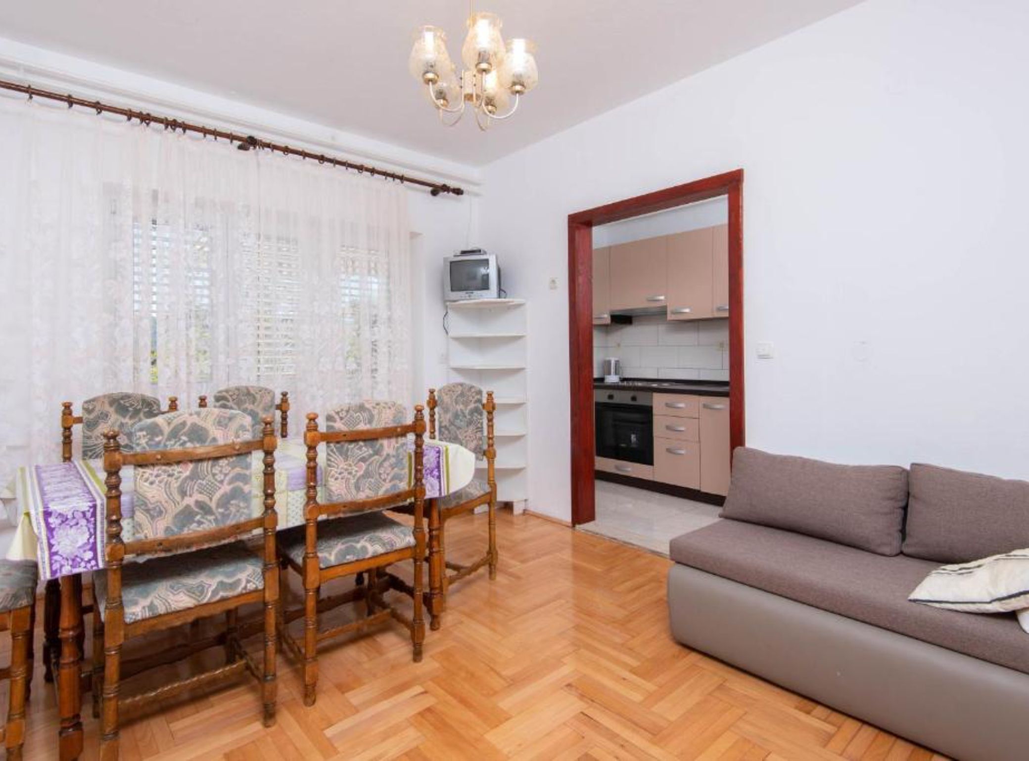 A living room with a sofa, a dining table with chairs, a kitchen area, and a chandelier.