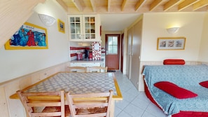 A room with a dining table, chairs, a bed with patterned bedding, a kitchen area with cabinets, and a door leading to another room.