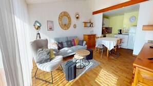 Ein Wohnzimmer mit einer Couch, Sesseln, einem Couchtisch und einem Essbereich mit Tisch und Stühlen. Im Hintergrund ist eine Küche erkennbar.