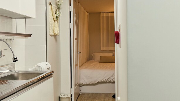 Ein enger Flur führt zu einem Schlafzimmer mit einem Bett, einer kleinen Küchenzeile mit Spüle und Schränken sowie einem Mülleimer.