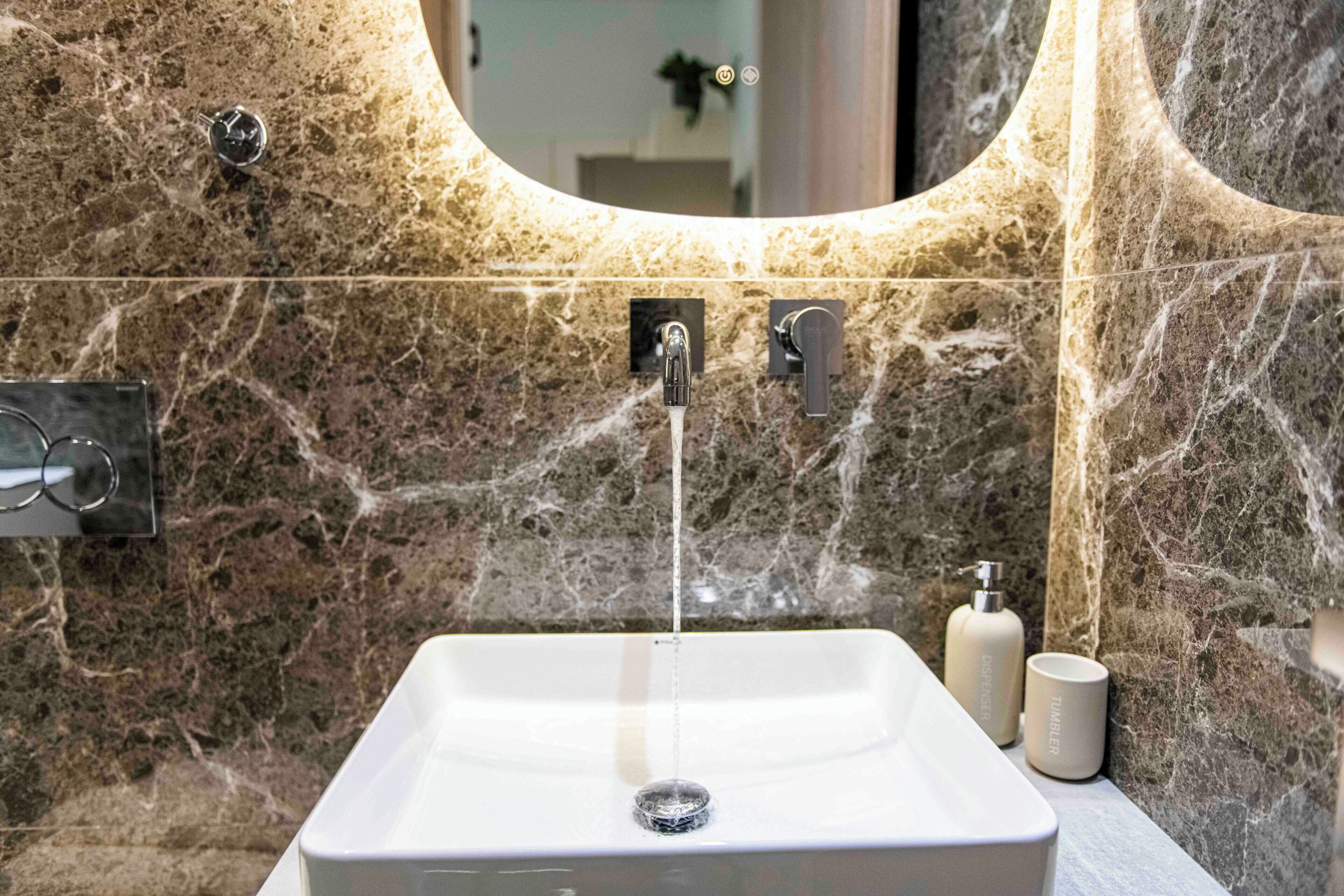 A bathroom with a marble sink, two faucets, a mirror, and a towel dispenser.