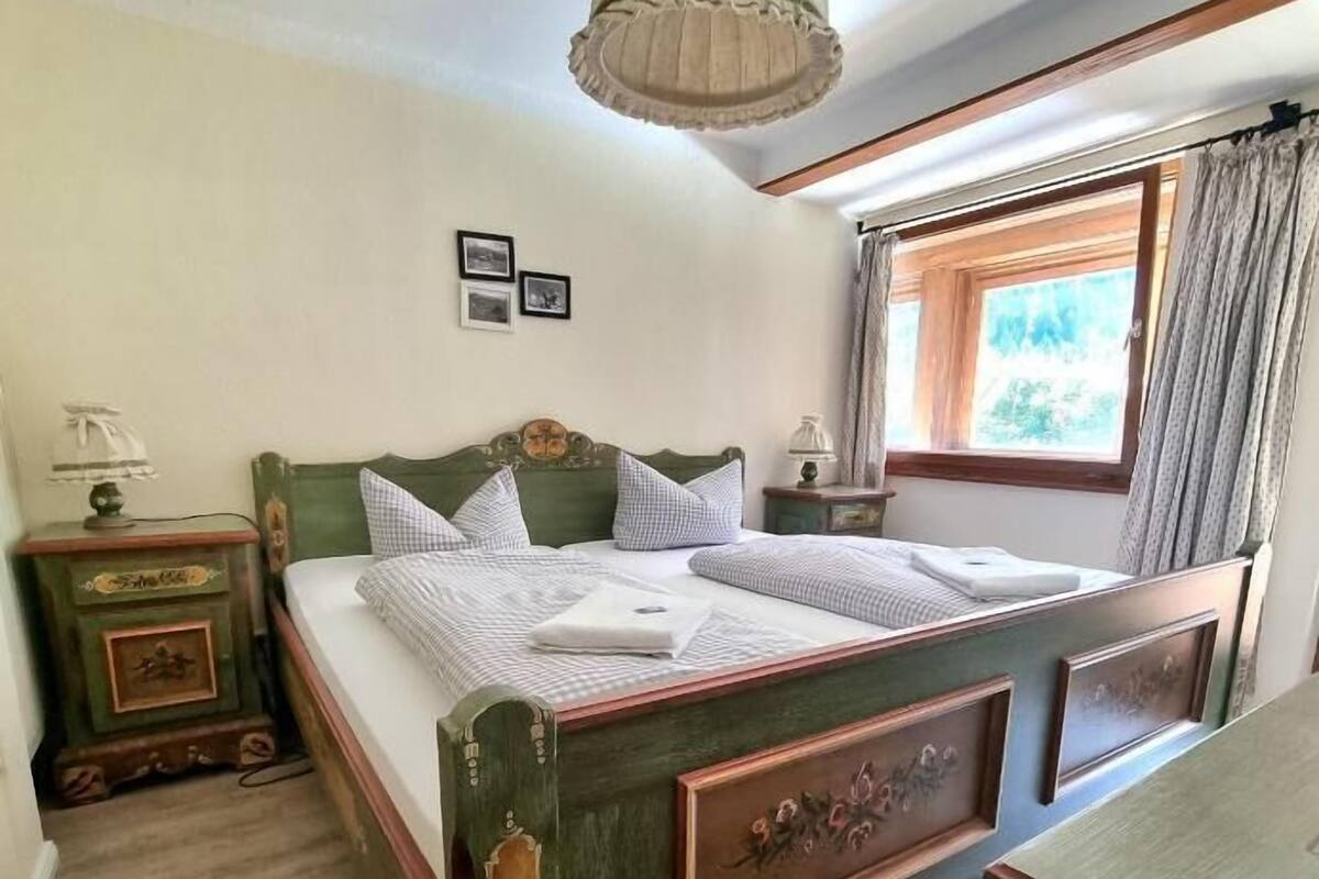 Ein Schlafzimmer mit einem hölzernen Bett, einem Nachttisch, einer Kommode, einem Fenster mit Vorhängen und einer Pendelleuchte.
