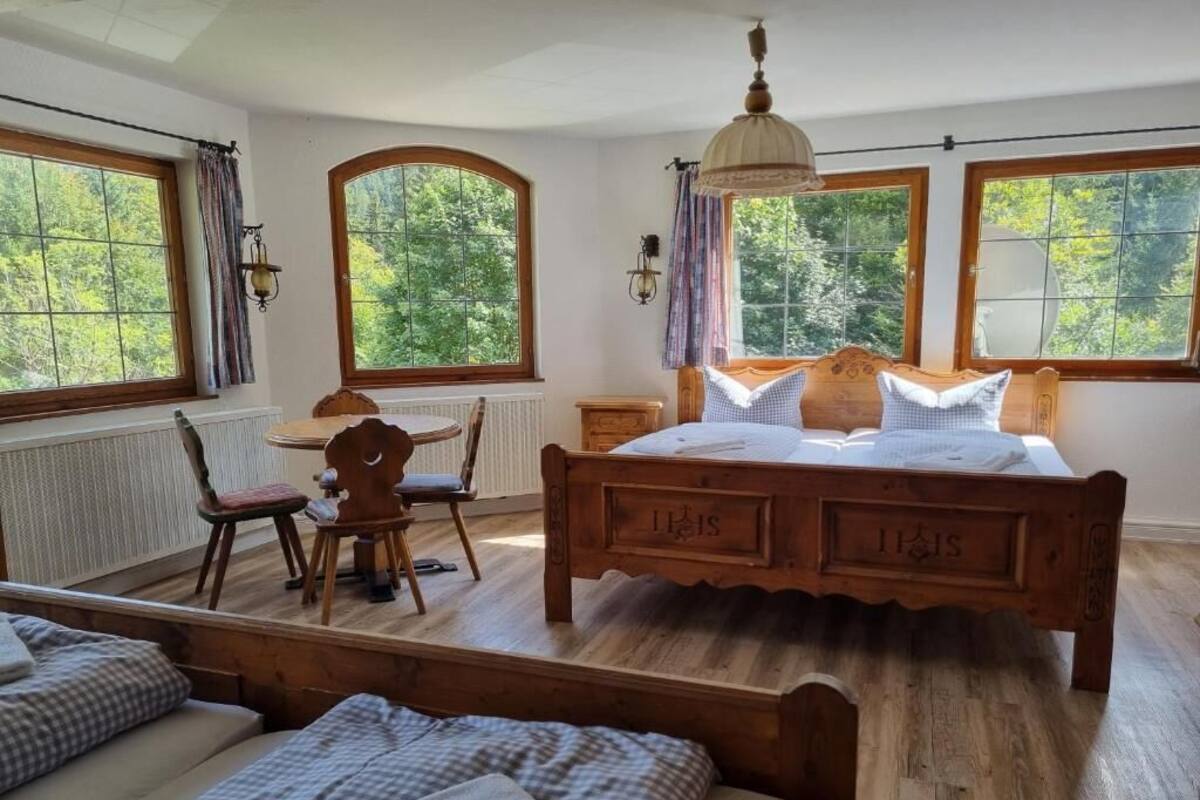 Ein Schlafzimmer mit einem hölzernen Bett, einem Sessel, einem Tisch und großen Fenstern mit Vorhängen.