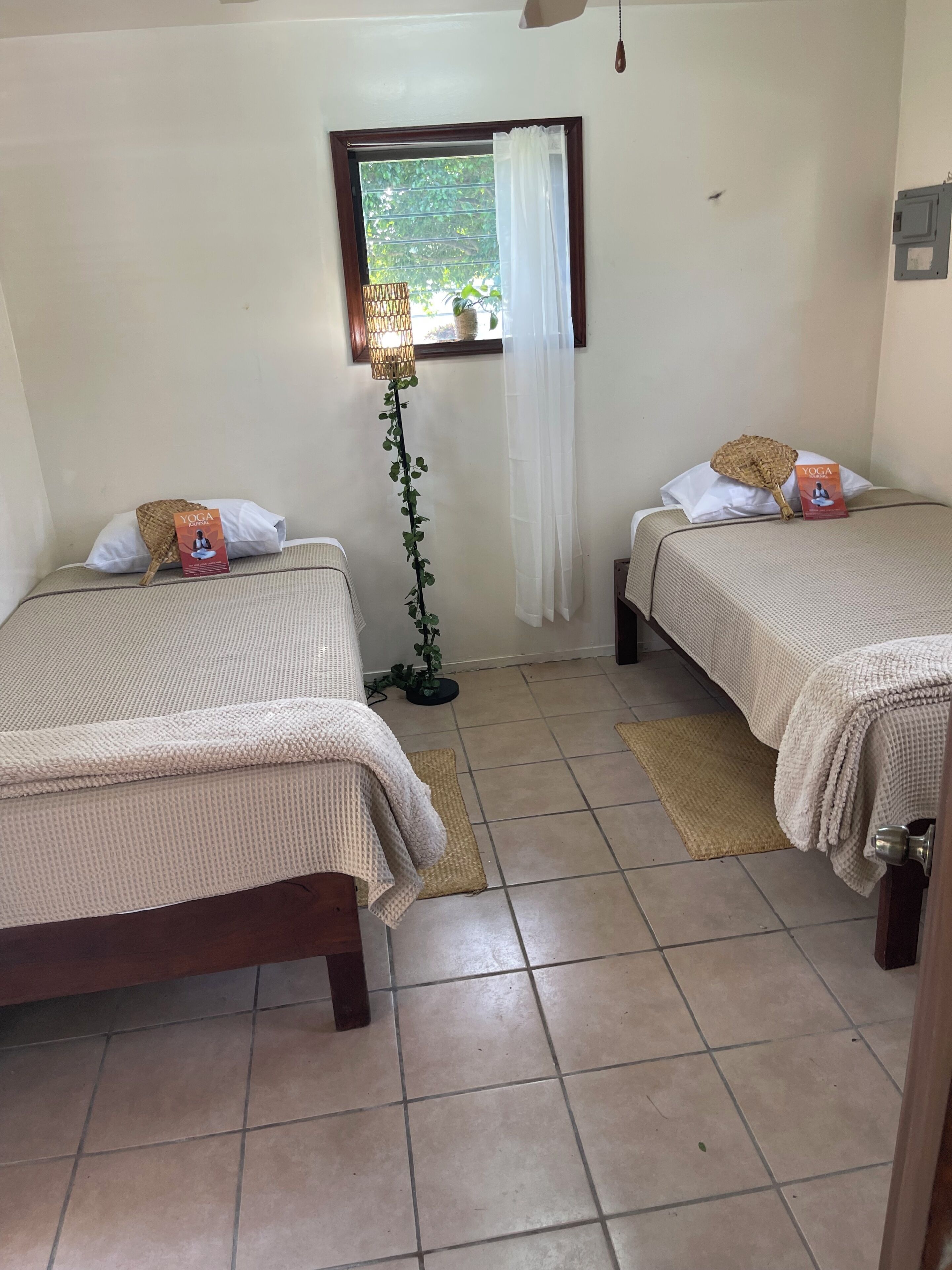 A room with two beds, a window with a curtain, a floor lamp, and a yoga book on one bed.