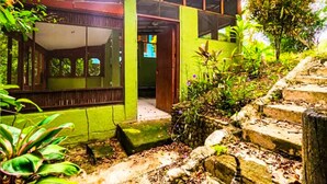 Een groen gebouw met een overdekte ingang, treden die erheen leiden en een tuin met planten en een stenen muur.