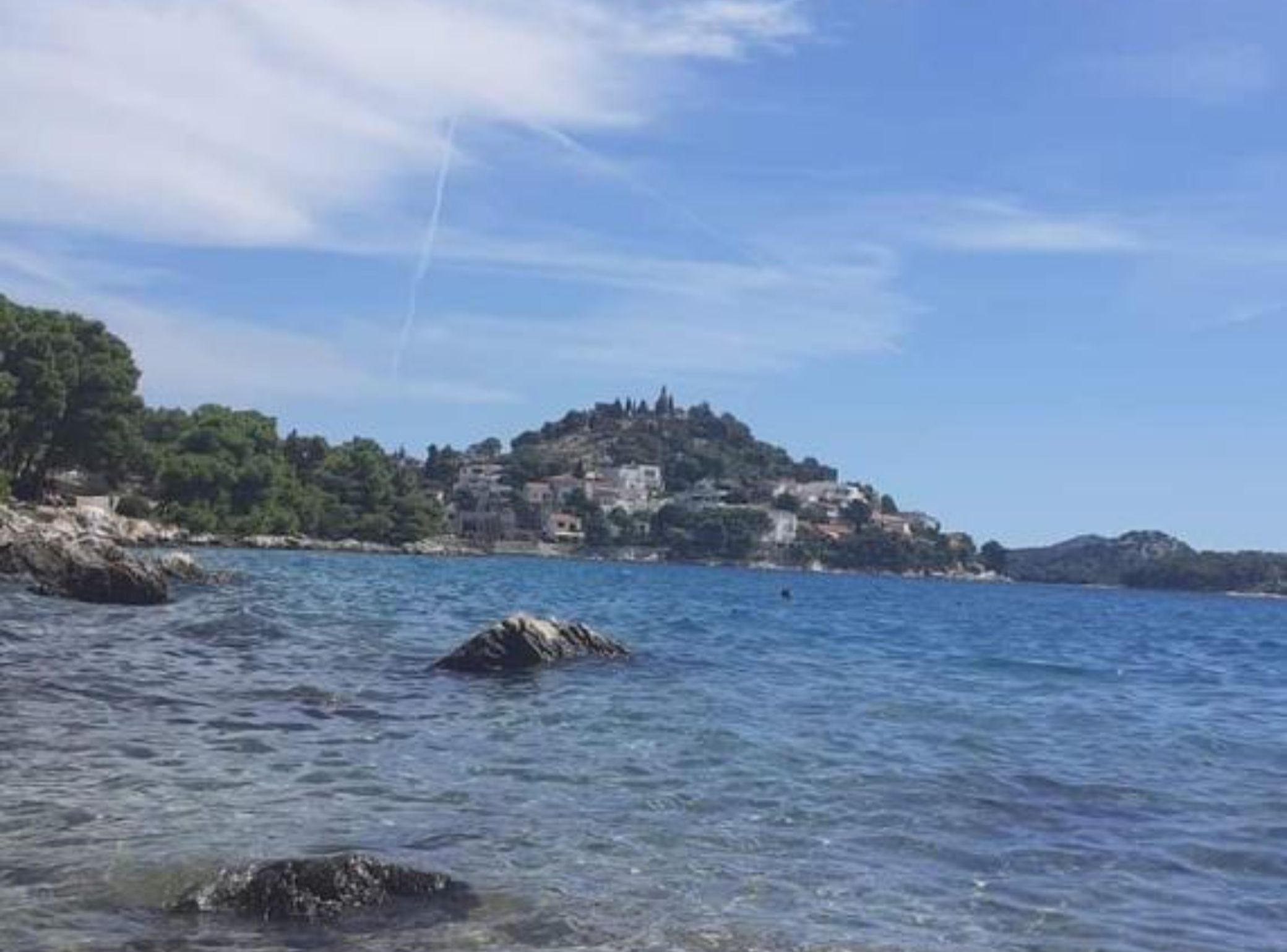 A coastal view with a rocky shoreline, clear blue water, and a hilltop with buildings.