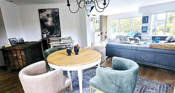 A modern living room with a round wooden table, two chairs, a blue sofa, and a large window offering a view of greenery.