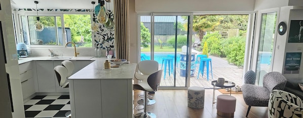 A modern kitchen with a large window, a dining area with blue chairs, and a view of a swimming pool. - Villa with south-facing swimming pool, a haven of peace on the outskirts of Vannes and the Gulf. (Ploeren)