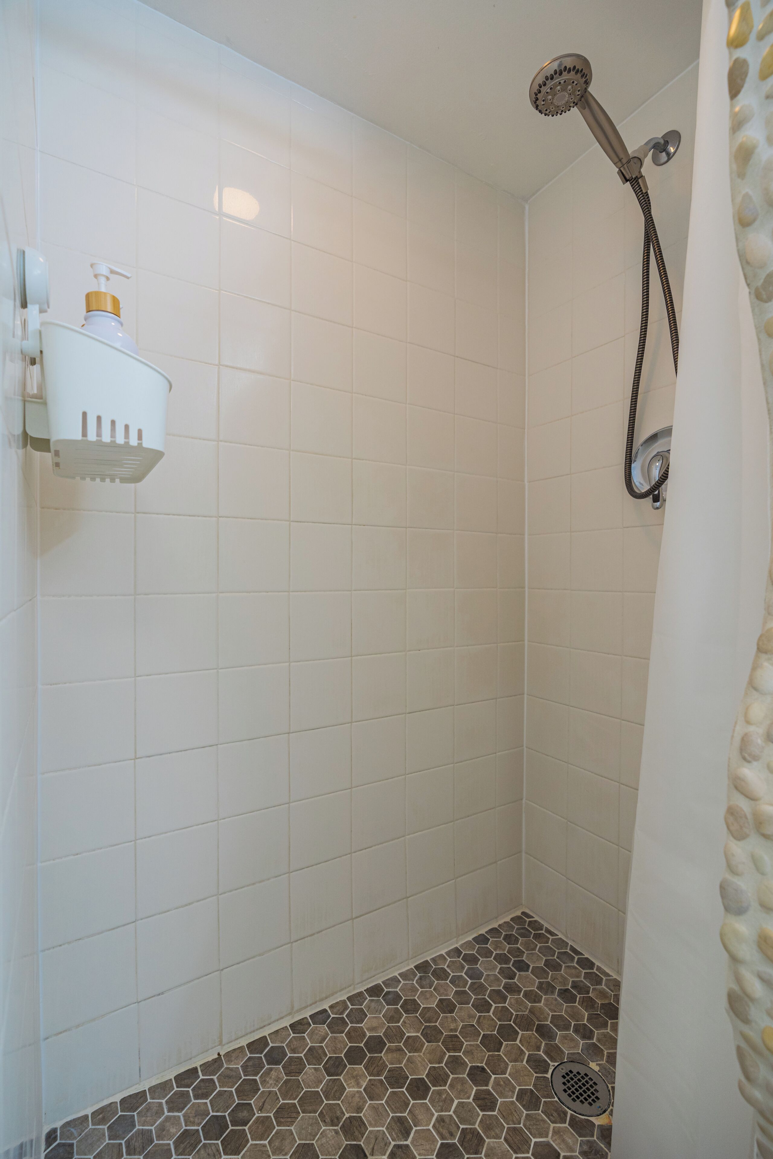 A shower with a white tiled wall, a showerhead, and a drain.