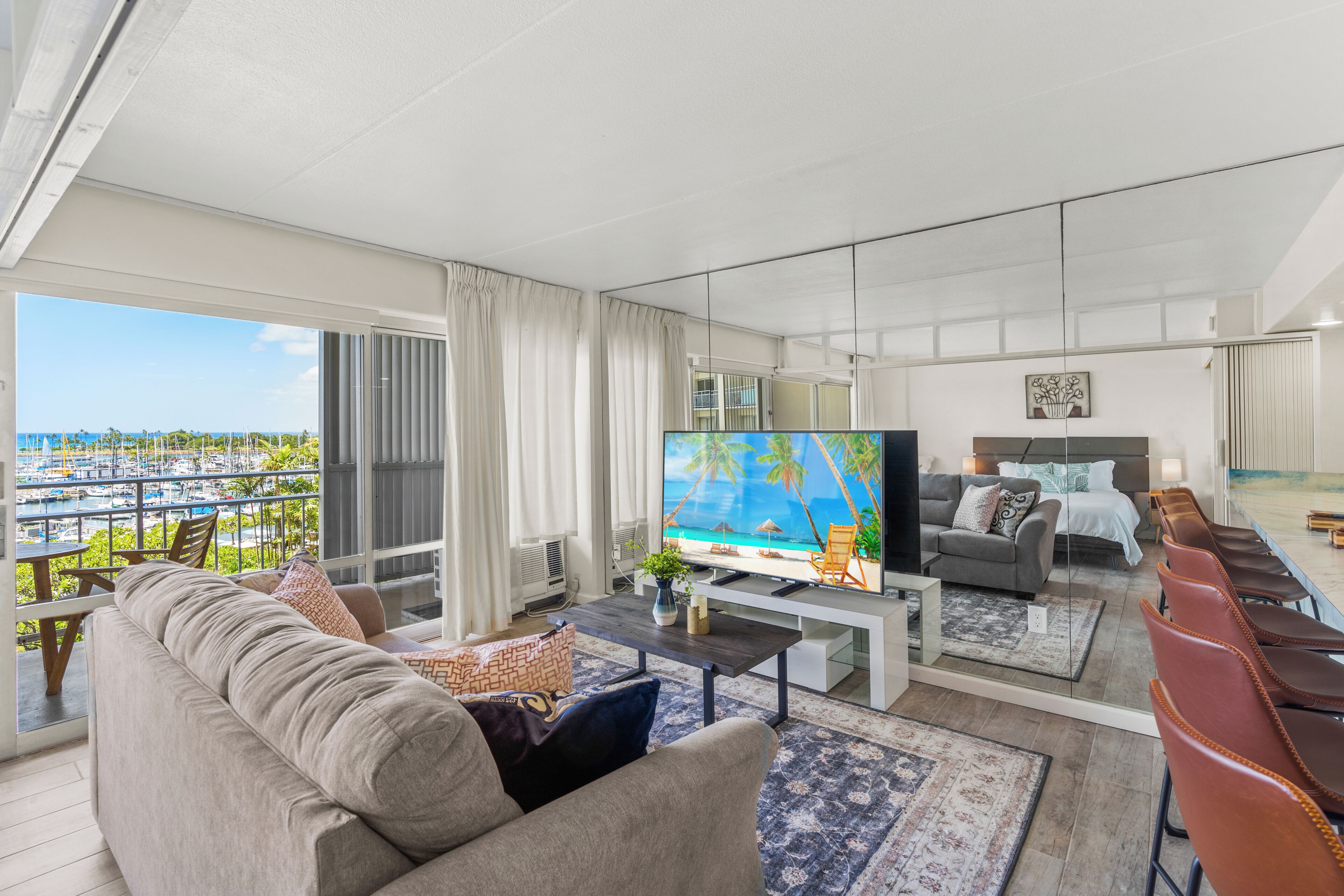 A spacious living room with a large TV, comfortable seating, and a balcony with a view of the ocean.