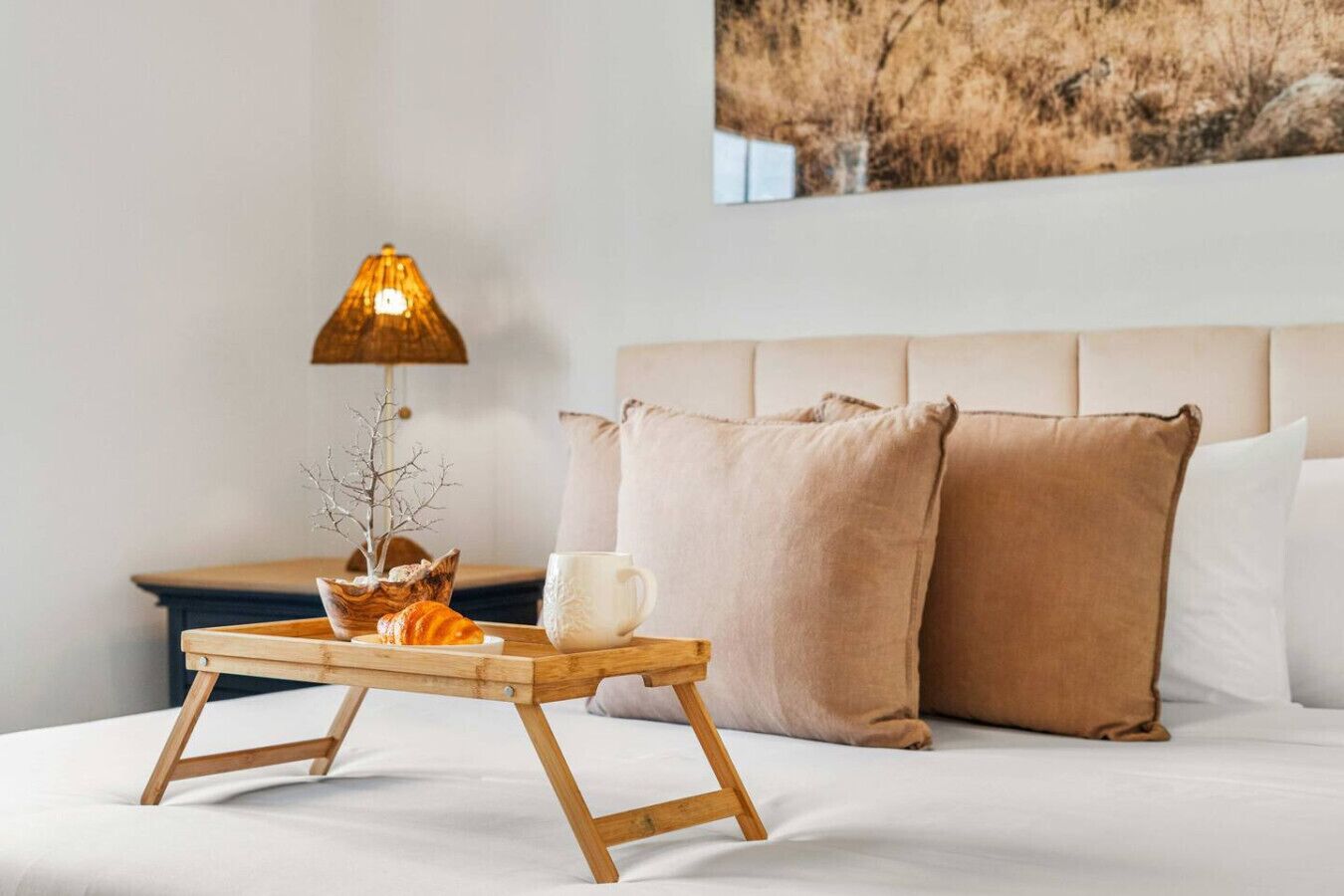 A bed with a wooden bedside table, a lamp, a small tree, a cup, and two pillows.