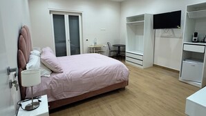 Ein Schlafzimmer mit Bett, Nachttisch, Lampe, Kleiderschrank, Fernseher und Schreibtisch mit Stuhl.