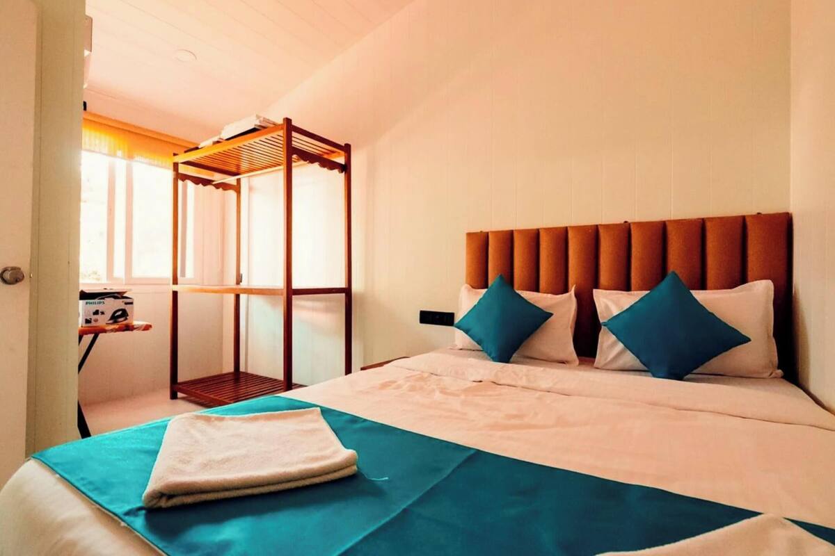 Quarto de hotel com uma cama, uma janela, uma cadeira de madeira e uma mesinha com uma luminária.