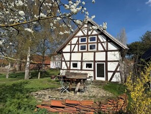 Una casa tradicional con entramado de madera, con una mesa y sillas de madera en primer plano, rodeada por un jardín con árboles en flor.