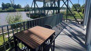 Eine Holzterrasse mit Tisch und Stühlen mit Blick auf einen Fluss und eine große Metallkonstruktion.