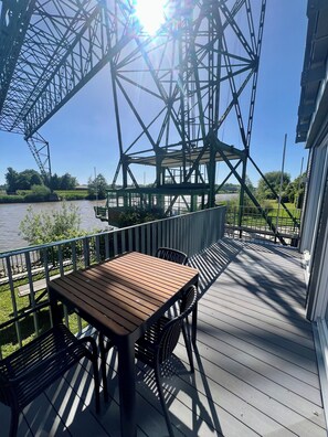 Terraza de madera con una mesa y sillas, con vista a un río y una gran estructura metálica.